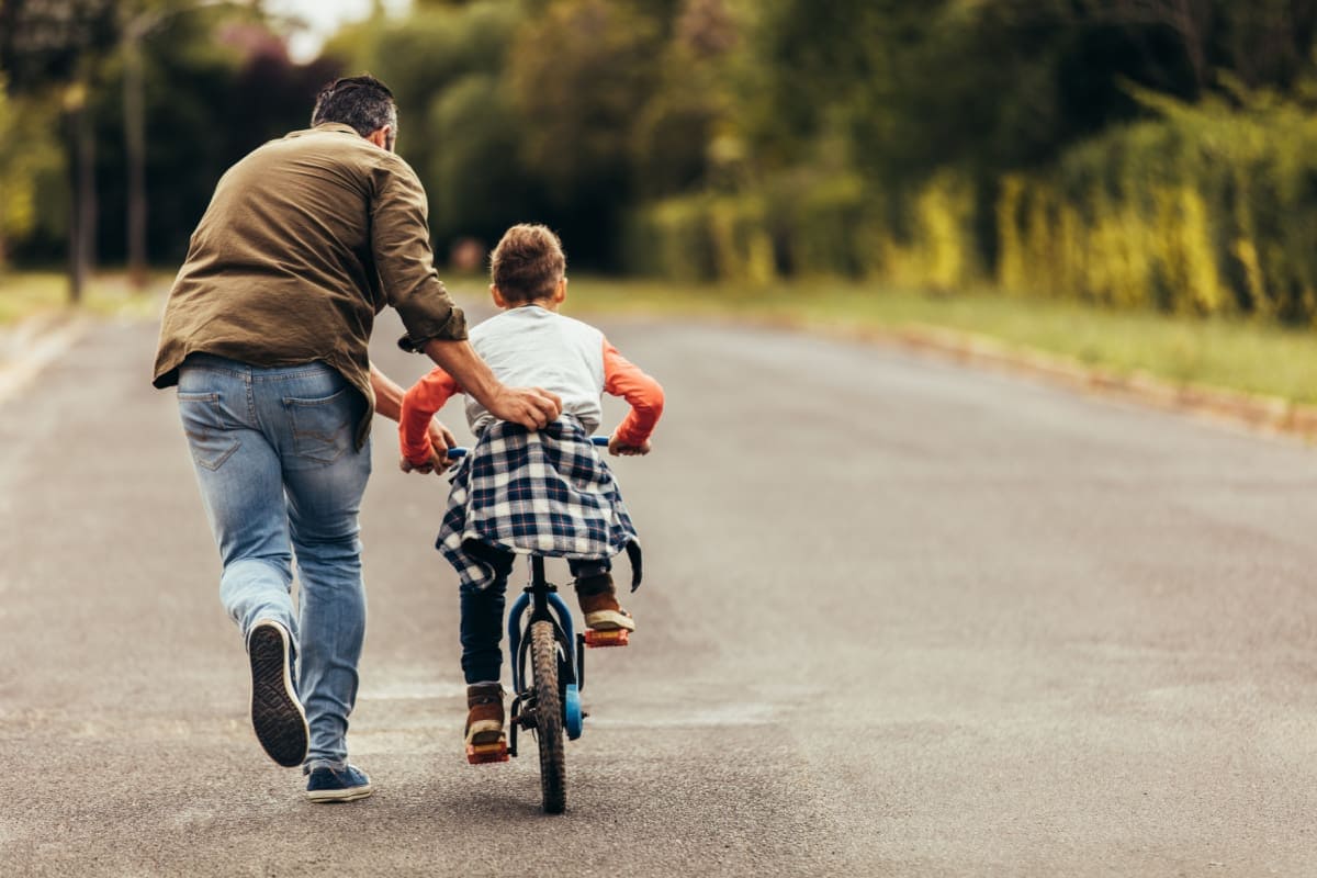 child riding bike along a road with adult pushing him along, helping him learn to ride a bicycle.